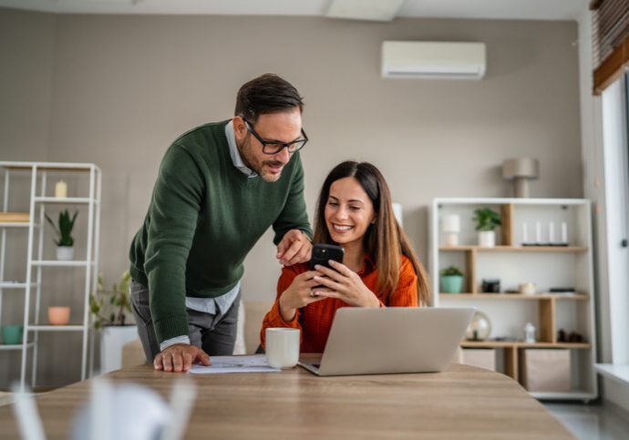 Smiling couple in a modern home office reviewing documents and managing finances together, using a laptop and a smart phone for budgeting and online banking